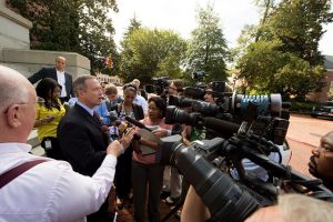 Man faces questions of a large number of reporters.